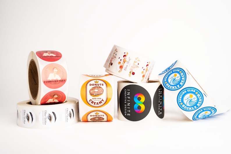 May include: Rolls of round stickers with various designs.  One roll features a donut with the text "Donuts Creamy Est'd 1995". Another roll features a rainbow infinity symbol with the text "Infinite Creative Design". A third roll features a blue wave with the text "Surfing Club Beckly Beach".