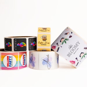 May include: Rolls of stickers with various designs, including a rainbow balloon party design, a black and white phoenix design, a watercolor design with the name "Amanda", a coffee shop design with a cartoon coffee cup, and a floral design with the text "BLOOMY BEAUTY BAR".