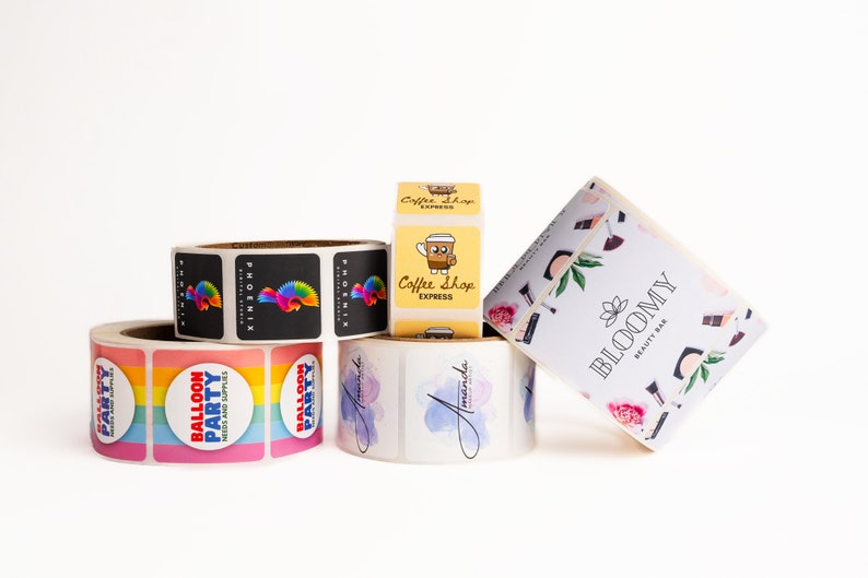 May include: Rolls of stickers with various designs, including a rainbow balloon party design, a black and white phoenix design, a watercolor design with the name "Amanda", a coffee shop design with a cartoon coffee cup, and a floral design with the text "BLOOMY BEAUTY BAR".