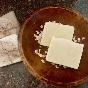 May include: Two bars of natural oatmeal soap in a wooden bowl with oats scattered around them. The soap is wrapped in brown paper with a twine bow and a label that reads "Natural Oatmeal Soap".