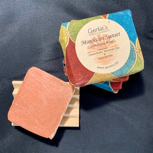 May include: A bar of Moroccan Sunset Detoxifying Soap by Gertie's Soaps & Sundries. The soap is made with cocoa butter, Moroccan clay, and essential oils. The soap is a light orange color and is sitting on a wooden soap dish.
