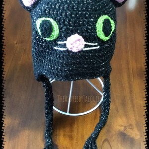 Puede incluir: Sombrero de gato negro de ganchillo con ojos verdes, orejas rosas y nariz rosa. El sombrero tiene largas borlas negras.