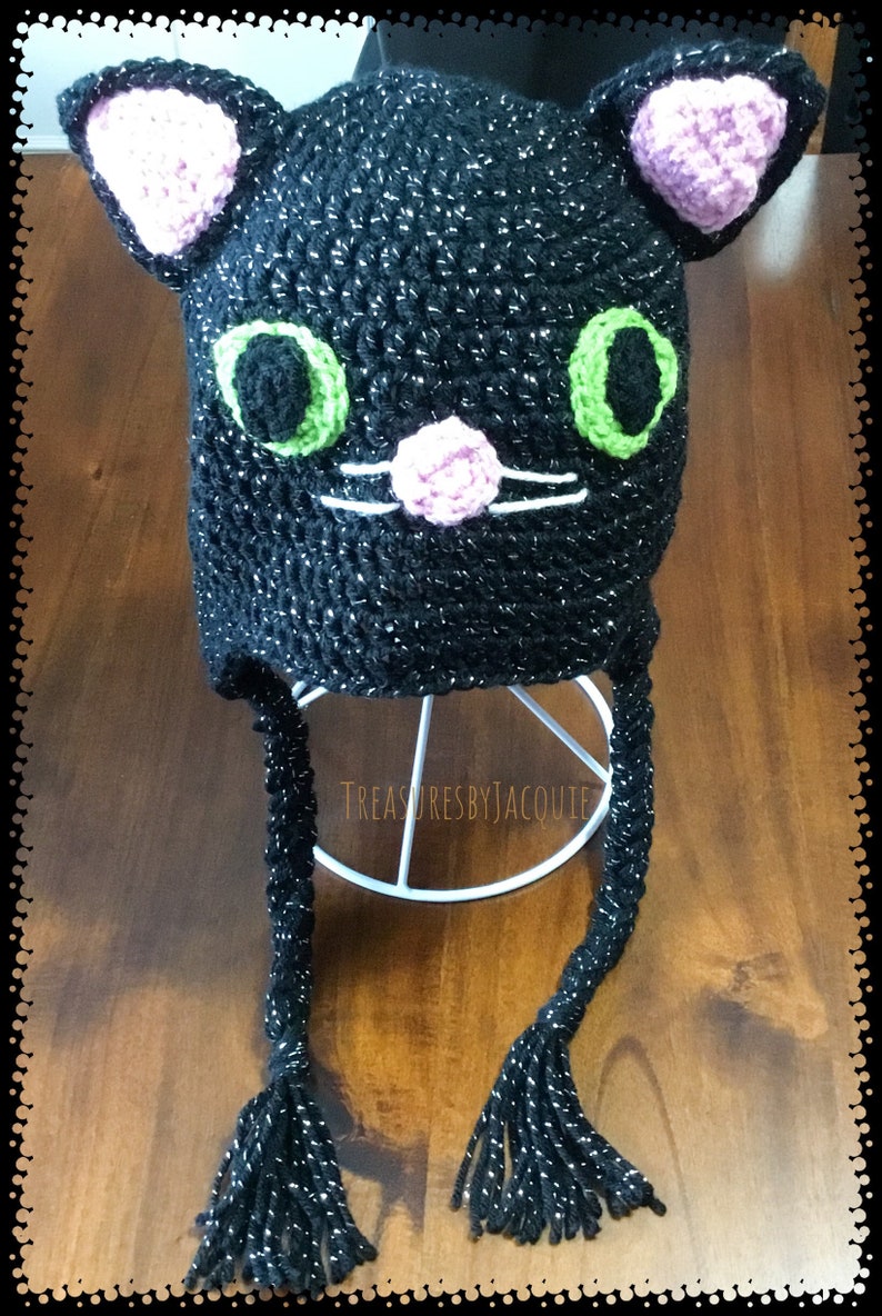 Puede incluir: Sombrero de gato negro de ganchillo con ojos verdes, orejas rosas y nariz rosa. El sombrero tiene largas borlas negras.