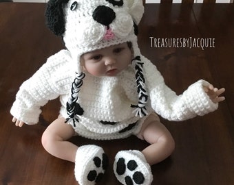 May include: A white crocheted baby outfit with black accents, featuring a dalmatian dog design. The outfit includes a hat, sweater, and booties. The hat has a black and white braided tail.