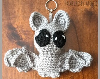 May include: A gray crocheted bat with black eyes and a silver key ring attached. The bat is on a wooden surface.