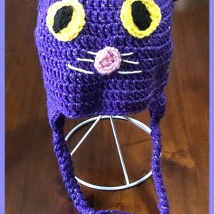 Puede incluir: Sombrero de gato de ganchillo morado con ojos amarillos y negros, nariz rosa y bigotes blancos. El sombrero tiene largas borlas moradas.