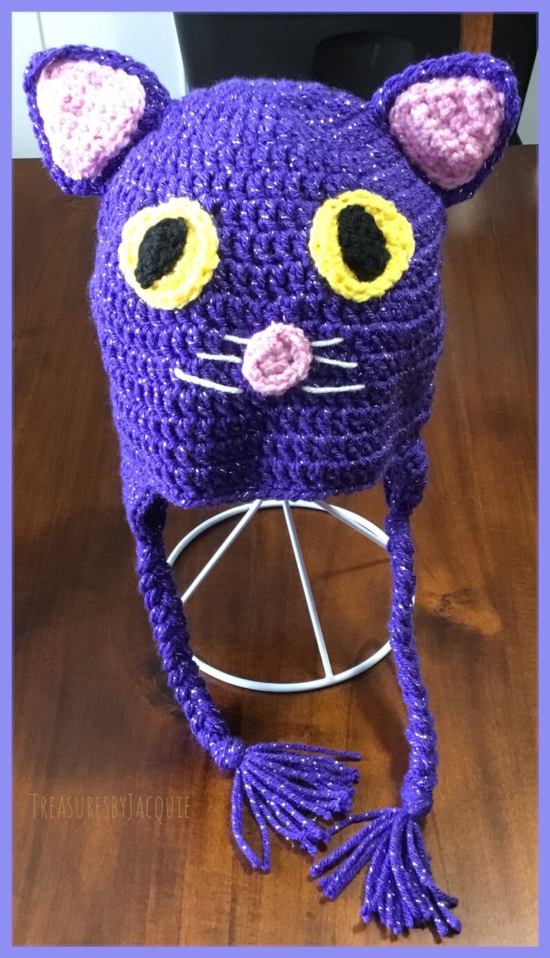 Puede incluir: Sombrero de gato de ganchillo morado con ojos amarillos y negros, nariz rosa y bigotes blancos. El sombrero tiene largas borlas moradas.