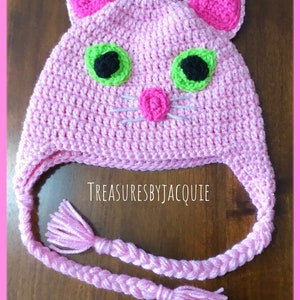 Puede incluir: Sombrero de gato de crochet rosa con ojos verdes, pupilas negras y una nariz rosa. El sombrero tiene una larga correa trenzada rosa con borlas en los extremos. El sombrero est&aacute; sobre una superficie de madera. TreasuresByJacquie est&aacute; escrito en el sombrero.