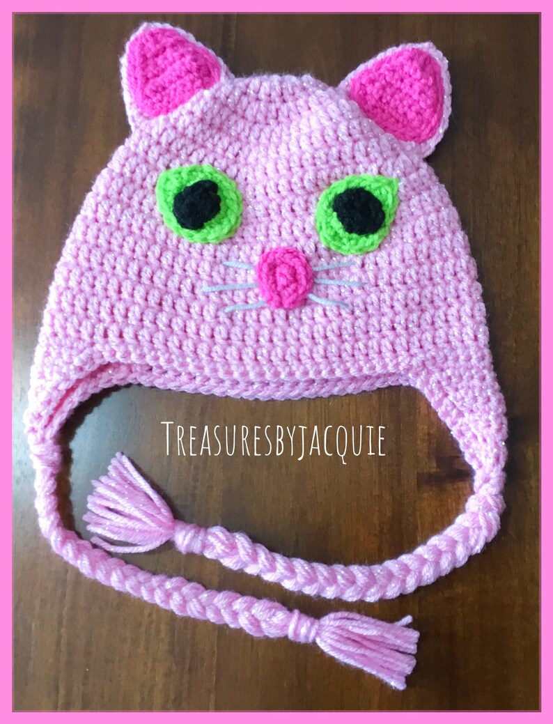 Puede incluir: Sombrero de gato de crochet rosa con ojos verdes, pupilas negras y una nariz rosa. El sombrero tiene una larga correa trenzada rosa con borlas en los extremos. El sombrero est&aacute; sobre una superficie de madera. TreasuresByJacquie est&aacute; escrito en el sombrero.