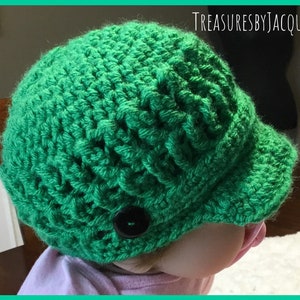Puede incluir: Un gorro verde tejido a ganchillo con una pequeña visera y un botón negro. El gorro está hecho a mano y tiene un patrón texturizado. El texto "TREASURESBYJACQUIE" es visible en la esquina superior derecha.