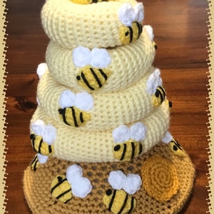 May include: A crocheted beehive stacking toy with a yellow and brown honeycomb base. The toy is made up of five rings with crocheted bees on each ring. The top ring has a single bee with white wings.