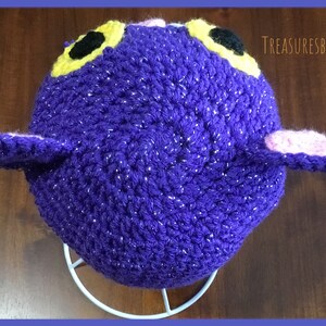 Puede incluir: Sombrero de crochet morado con orejas rosas y amarillas y ojos negros. El sombrero tiene una textura brillante. El sombrero est&aacute; en un soporte blanco.