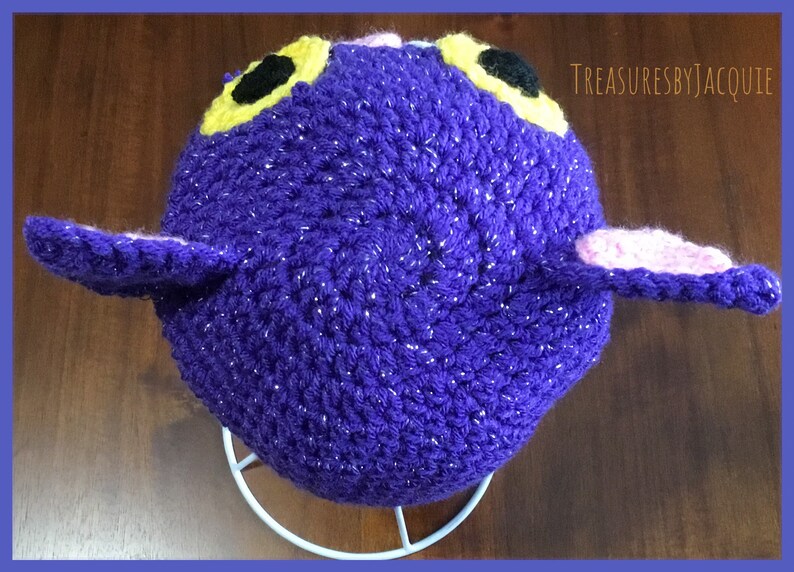 Puede incluir: Sombrero de crochet morado con orejas rosas y amarillas y ojos negros. El sombrero tiene una textura brillante. El sombrero est&aacute; en un soporte blanco.
