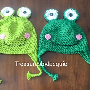 May include: Two green crocheted frog hats with white eyes and pink cheeks. The hats have long ties that can be tied under the chin.