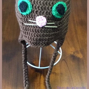 Puede incluir: Sombrero de gato de ganchillo marr&oacute;n con orejas rosas, ojos verdes y nariz rosa. El sombrero tiene dos largas borlas marrones.