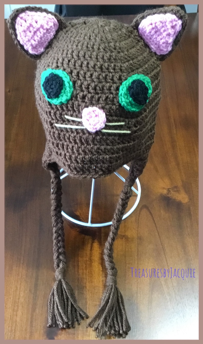Puede incluir: Sombrero de gato de ganchillo marr&oacute;n con orejas rosas, ojos verdes y nariz rosa. El sombrero tiene dos largas borlas marrones.