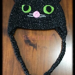 Puede incluir: Sombrero de gato negro de ganchillo con ojos verdes, orejas y nariz rosas y bigotes blancos. El sombrero tiene una larga borla negra.