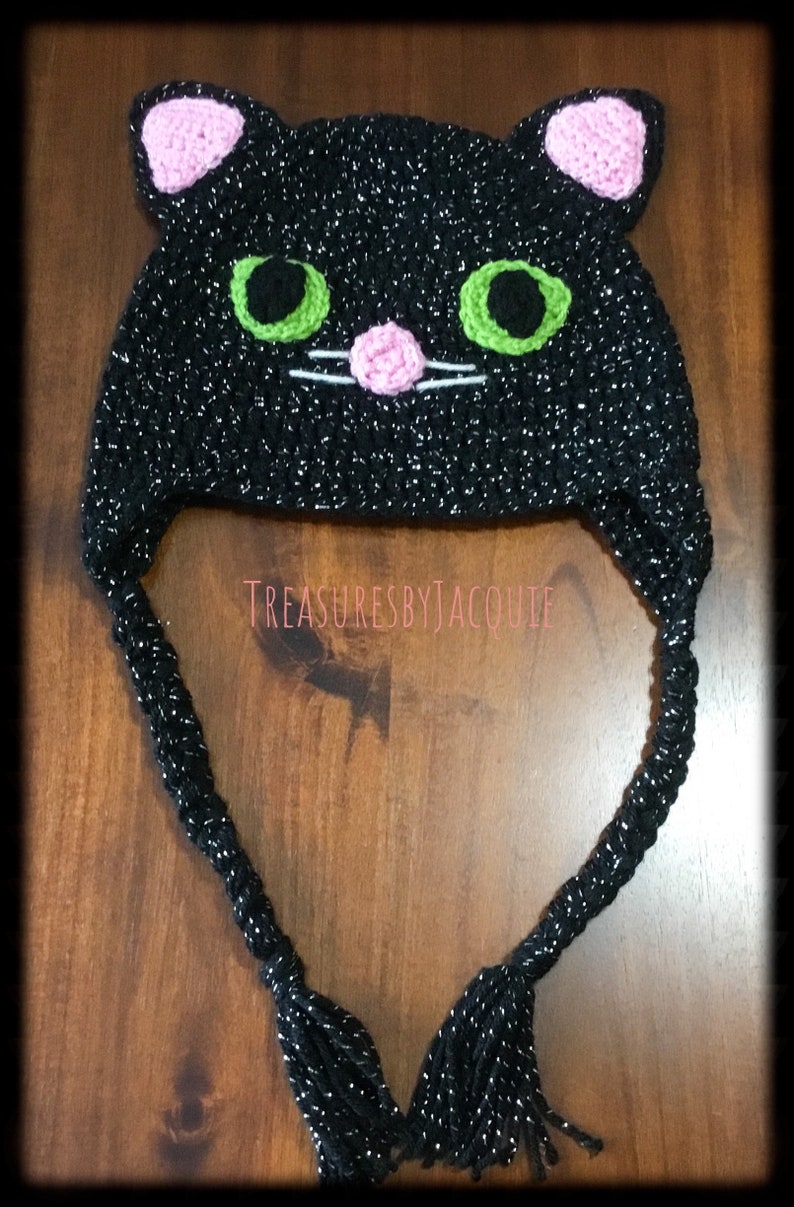 Puede incluir: Sombrero de gato negro de ganchillo con ojos verdes, orejas y nariz rosas y bigotes blancos. El sombrero tiene una larga borla negra.