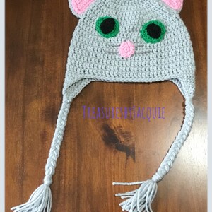Puede incluir: Sombrero de gato de crochet gris con orejas rosas, ojos verdes y una nariz rosa. El sombrero tiene trenzas grises largas con borlas.