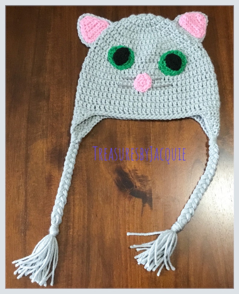 Puede incluir: Sombrero de gato de crochet gris con orejas rosas, ojos verdes y una nariz rosa. El sombrero tiene trenzas grises largas con borlas.