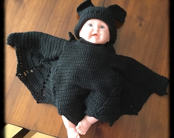 May include: A black crochet baby bat costume, including a hat with bat ears and a bodysuit with attached wings. The costume is designed to fit a baby. The baby is lying on a wooden surface.