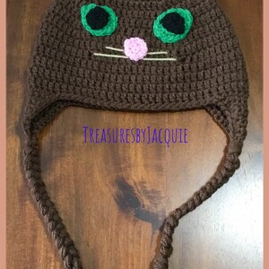 Puede incluir: Sombrero de gato de ganchillo marr&oacute;n con orejas rosas, ojos verdes y nariz rosa. El sombrero tiene largas borlas marrones.