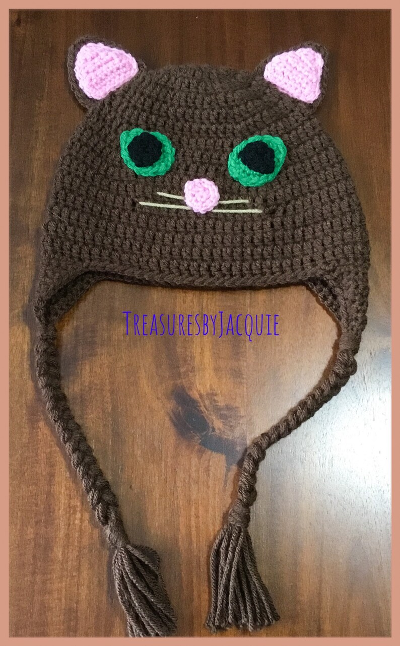 Puede incluir: Sombrero de gato de ganchillo marr&oacute;n con orejas rosas, ojos verdes y nariz rosa. El sombrero tiene largas borlas marrones.