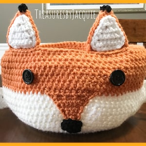 Puede incluir: Una cesta de crochet con forma de zorro hecha con hilo naranja y blanco, ojos de botón negros y una nariz negra. La cesta está sentada sobre una mesa de madera.
