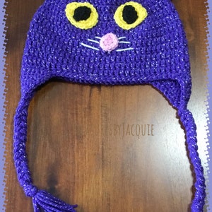 Puede incluir: Sombrero de gato de ganchillo morado con hilo brillante y bigotes blancos. El sombrero tiene orejas rosas y ojos amarillos.
