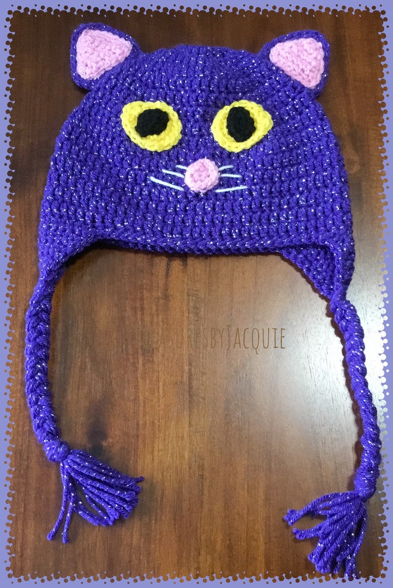 Puede incluir: Sombrero de gato de ganchillo morado con hilo brillante y bigotes blancos. El sombrero tiene orejas rosas y ojos amarillos.