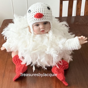 Puede incluir: Un bebé con un disfraz de pollo de crochet blanco hecho a mano, con un gorro a juego, botines rojos de crochet y detalles de plumas. El gorro tiene ojos de botón negros y un pico rojo. El texto "TreasuresbyJacquie" es visible.