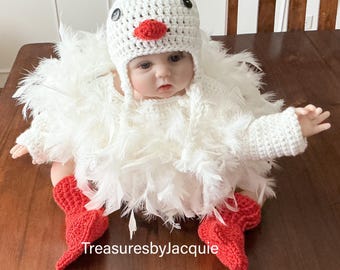 May include: A baby in a handmade white crochet chicken costume, including a hat, red crochet booties, and feather detailing. The hat features black button eyes and a red beak. The text "TreasuresbyJacquie" is visible.