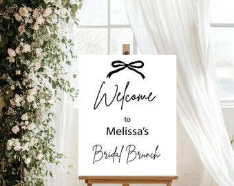 Pode incluir: Uma placa branca em um cavalete de madeira com o texto "Welcome to Melissa's Bridal Brunch" em letras pretas. Um laço preto no topo. A placa está em frente a uma janela com cortinas brancas e um arco floral.