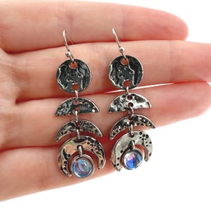 May include: Silver-toned dangle earrings with a celestial theme. Each earring features a circular top, a crescent-shaped middle, and a bottom crescent holding a blue iridescent bead. The earrings are designed with a textured, hammered metal finish.