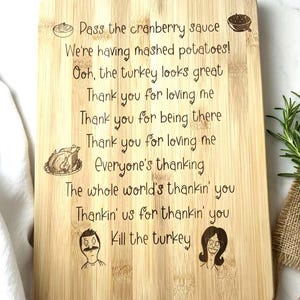 May include: A rectangular bamboo cutting board with engraved text and illustrations. The text includes phrases like "Pass the cranberry sauce" and "Kill the turkey." The board features illustrations of food and cartoon characters.