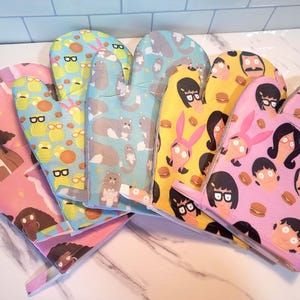 May include: A collection of colorful oven mitts with various cartoon character designs. The mitts feature characters with glasses, cats, and burger designs. The mitts are in shades of pink, yellow, blue, and green.