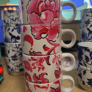 May include: Stacked white ceramic mugs with pink floral designs. Each mug has a rounded handle and a different floral pattern. Other mugs with blue and white floral patterns are visible in the background.