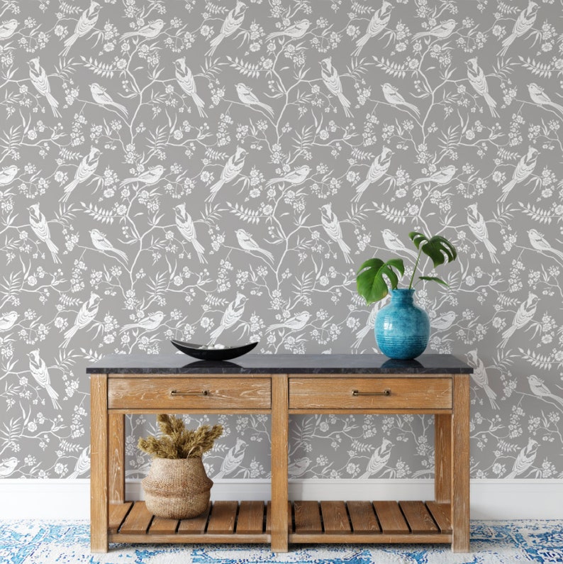 May include: A gray wallpaper with a white bird and floral pattern. A wooden console table with two drawers and a shelf sits in front of the wallpaper. A blue vase with a green plant sits on the table.