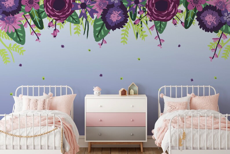 May include: Two white twin beds with pink bedding and a white dresser with pink, grey, and white drawers. The wall behind the beds is painted a light purple and features a floral mural with purple, pink, and green flowers.
