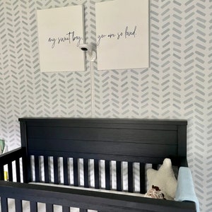 May include: A black crib with a slatted side and headboard is in a nursery. Two white framed canvases hang above the crib, each with black script. The canvases read "my sweet boy" and "you are so loved." A baby monitor is mounted between the canvases.