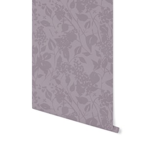 May include: A roll of wallpaper featuring a repeating floral pattern in shades of lavender and gray. The design includes stylized flowers, leaves, and stems, creating a soft, botanical aesthetic. The wallpaper is partially unrolled, revealing the pattern.