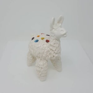 Puede incluir: Figura de llama de cer&aacute;mica blanca con detalles de pintura de colores. La llama tiene una superficie texturizada y est&aacute; de pie sobre una superficie blanca.