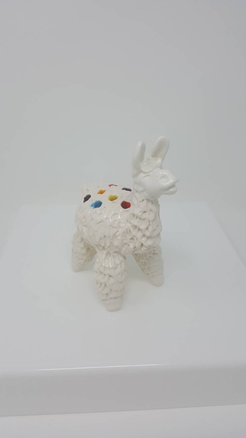 Puede incluir: Figura de llama de cer&aacute;mica blanca con detalles de pintura de colores. La llama tiene una superficie texturizada y est&aacute; de pie sobre una superficie blanca.