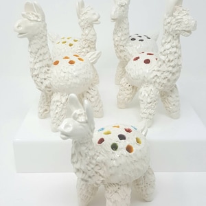 Puede incluir: Cinco esculturas de llama de cer&aacute;mica blanca con puntos de colores en la espalda. Las llamas est&aacute;n de pie sobre una superficie blanca.