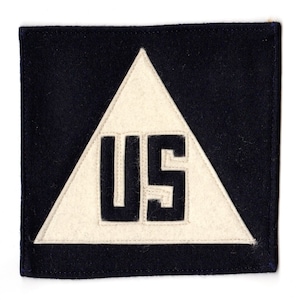 Può includere: Una toppa in feltro blu navy con un triangolo bianco che contiene le lettere "US" in nero.