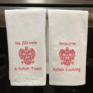 May include: Two white kitchen towels with red embroidered Polish sayings and the Polish coat of arms. The first towel says "Na Zdrowie A Polish Toast" and the second towel says "Smacze Polish Cooking".