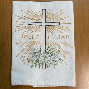K&ouml;nnte beinhalten: Wei&szlig;es K&uuml;chentuch mit einem gestickten Kreuz, Lilien und den Worten "Halleluja" in goldener Farbe.