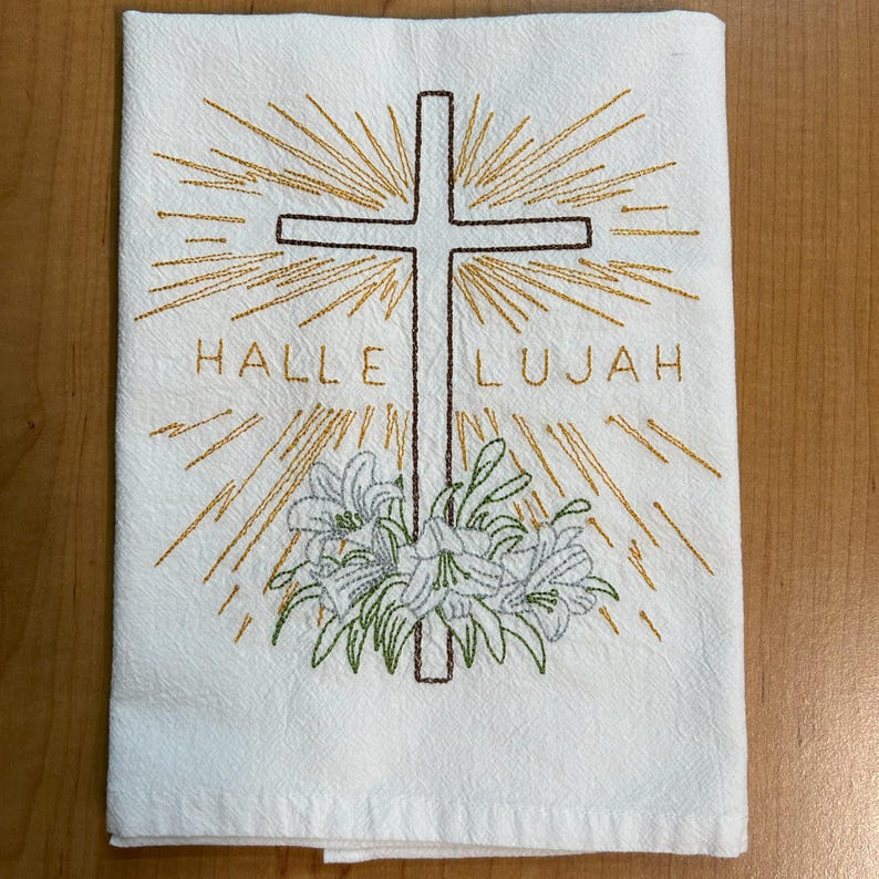 K&ouml;nnte beinhalten: Wei&szlig;es K&uuml;chentuch mit einem gestickten Kreuz, Lilien und den Worten "Halleluja" in goldener Farbe.