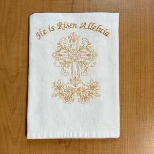 K&ouml;nnte beinhalten: Wei&szlig;es K&uuml;chentuch mit goldener Stickerei eines Kreuzes und Lilien. Der Text "He is Risen Alleluia" ist &uuml;ber dem Kreuz gestickt.