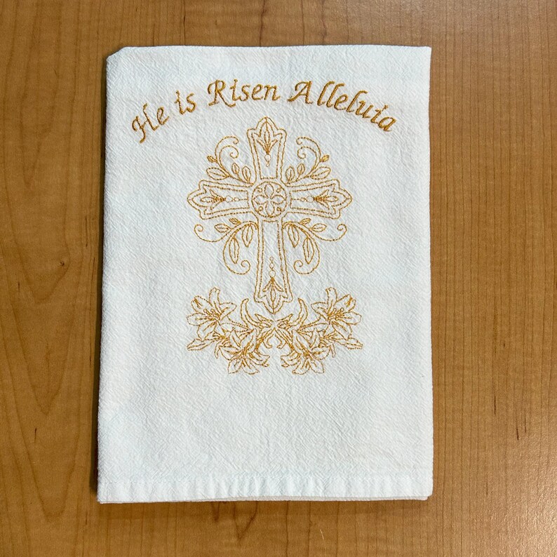 K&ouml;nnte beinhalten: Wei&szlig;es K&uuml;chentuch mit goldener Stickerei eines Kreuzes und Lilien. Der Text "He is Risen Alleluia" ist &uuml;ber dem Kreuz gestickt.